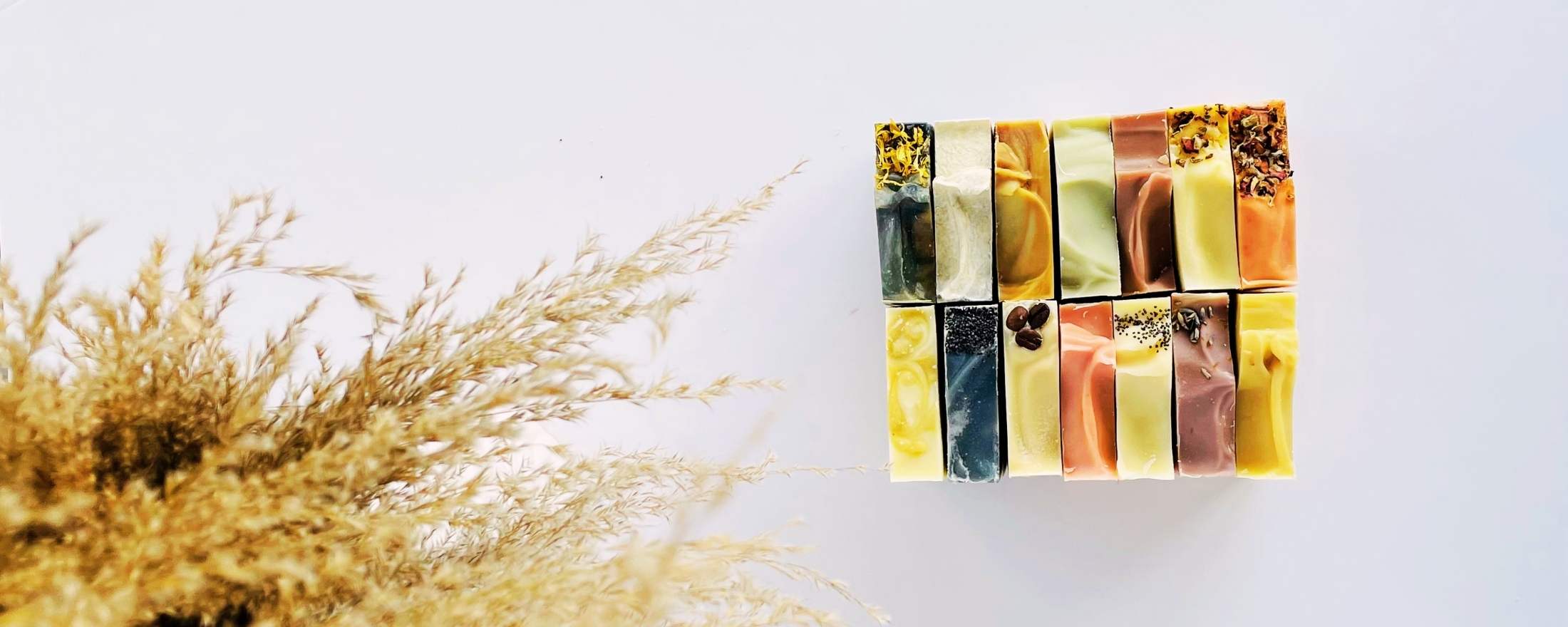 A top view of different bars of soap, each with unique colors and textures, arranged neatly in a grid pattern. The soaps are shown on a white surface with a text overlay that reads, "Healthy skin starts from within - prioritize nourishing your skin with high-quality natural skincare products, just as you prioritize your body with a balanced diet." There is also a button displayed that leads to a page where all products are available for shopping.